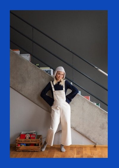 Person in white overalls and dark shirt leaning against a stairway wearing a hat.