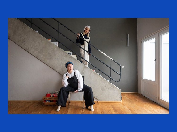 Two people wearing dark and light versions of the same overalls. One is walking down the stairs and the other is sitting in front of them.