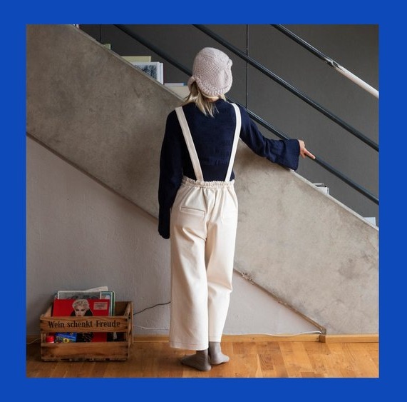 Back view of a person wearing cream overalls with straight straps and a dark shirt facing a wall.