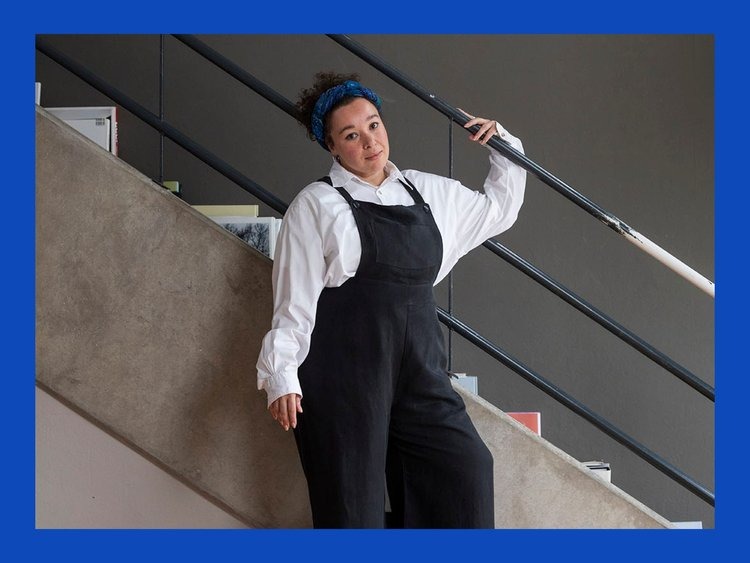 Person wearing overalls and a white shirt posing with one arm on a stair railing.