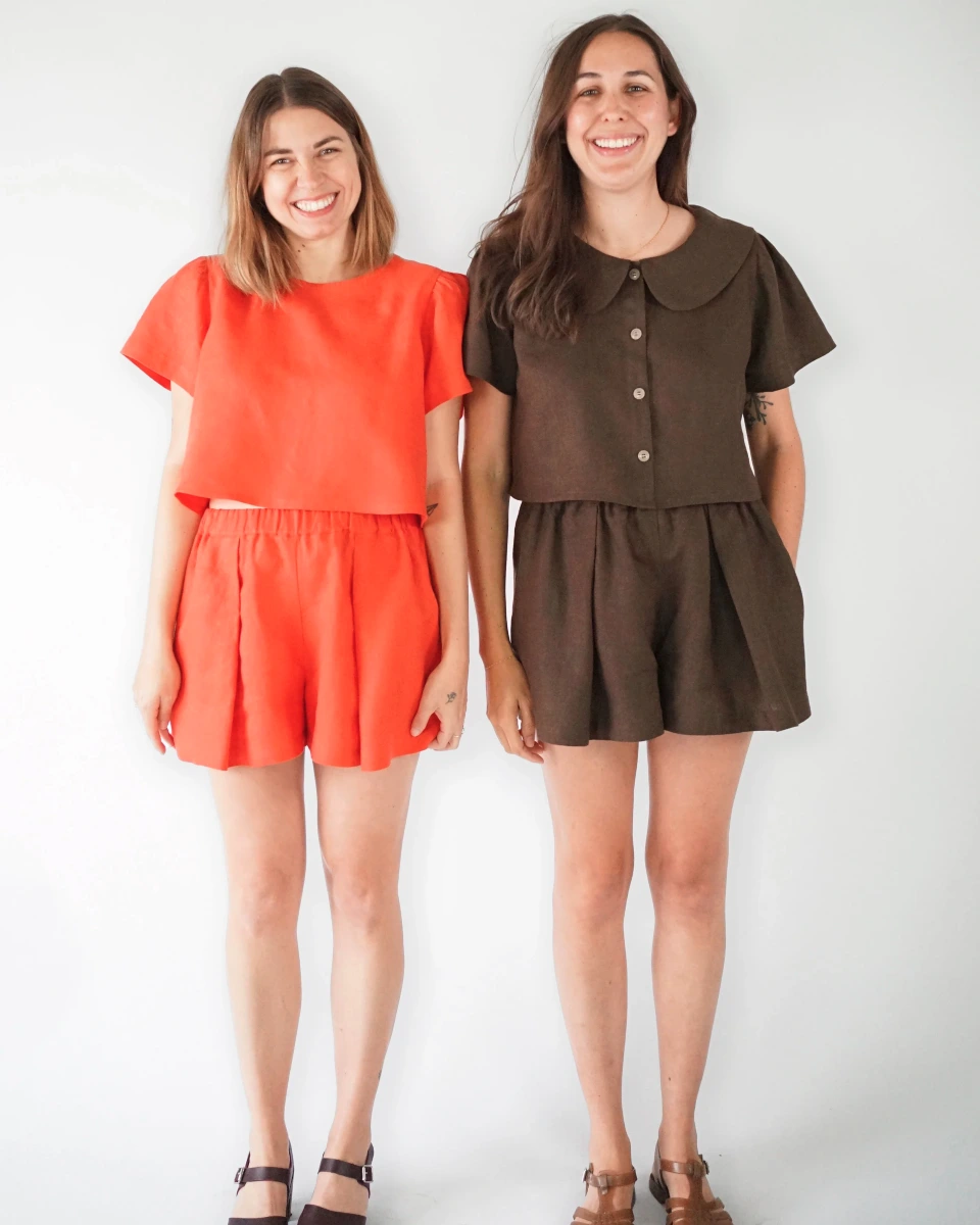 A bright red-orange short-sleeved top and shorts set (left) and a brown button-front blouse and shorts set (right) modeled on two smiling thin white women with blonde and brown hair.