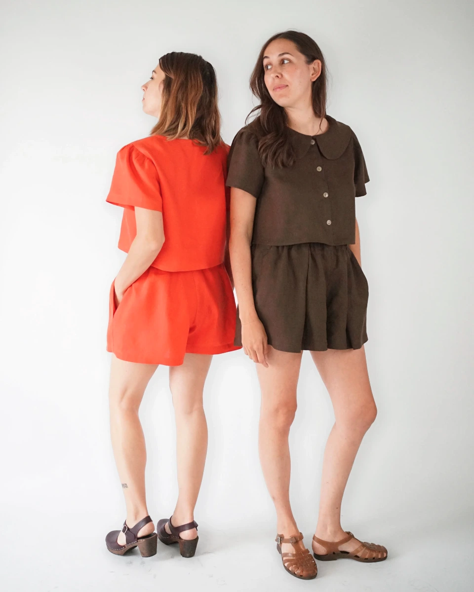 A bright red-orange short-sleeved top and shorts set seen from a rear three-quarter angle (left) and a brown button-front blouse and shorts set (right) seen from a front three-quarter angle modeled on two smiling thin white women with blonde and brown hair.