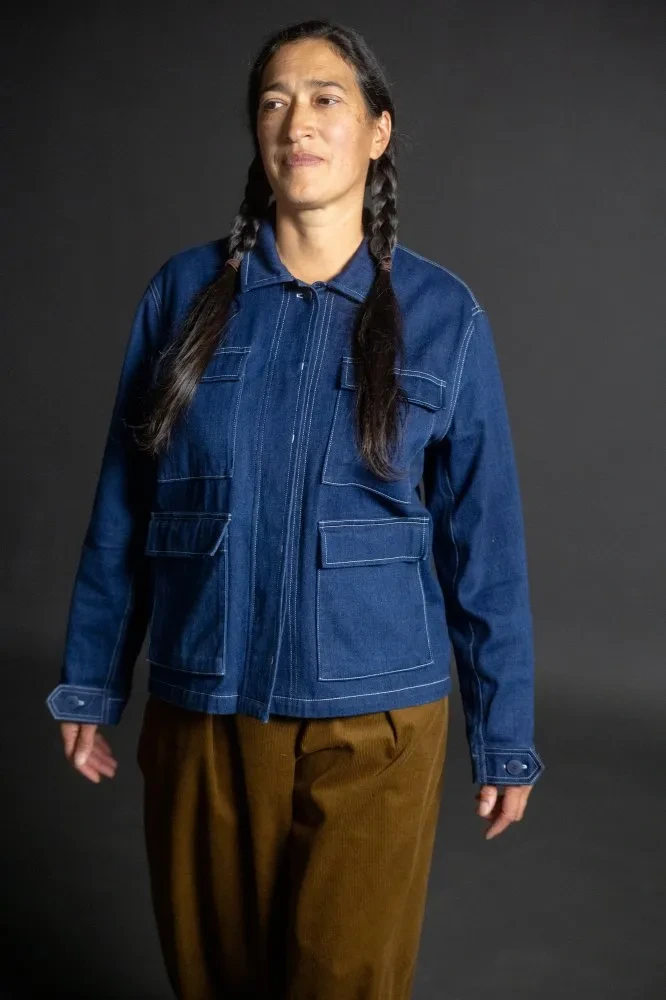 A dark indigo denim jacket with square elbow patches, square pockets with flaps, white contrast top-stitching, and a hidden button placket. It is modeled on a slim woman with brown hair in two braids.