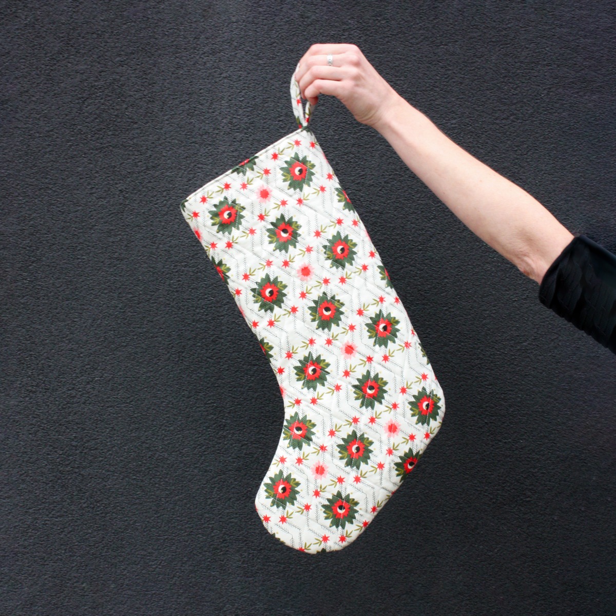 A white stocking with red and green abstract flowers held in front of a dark gray background.